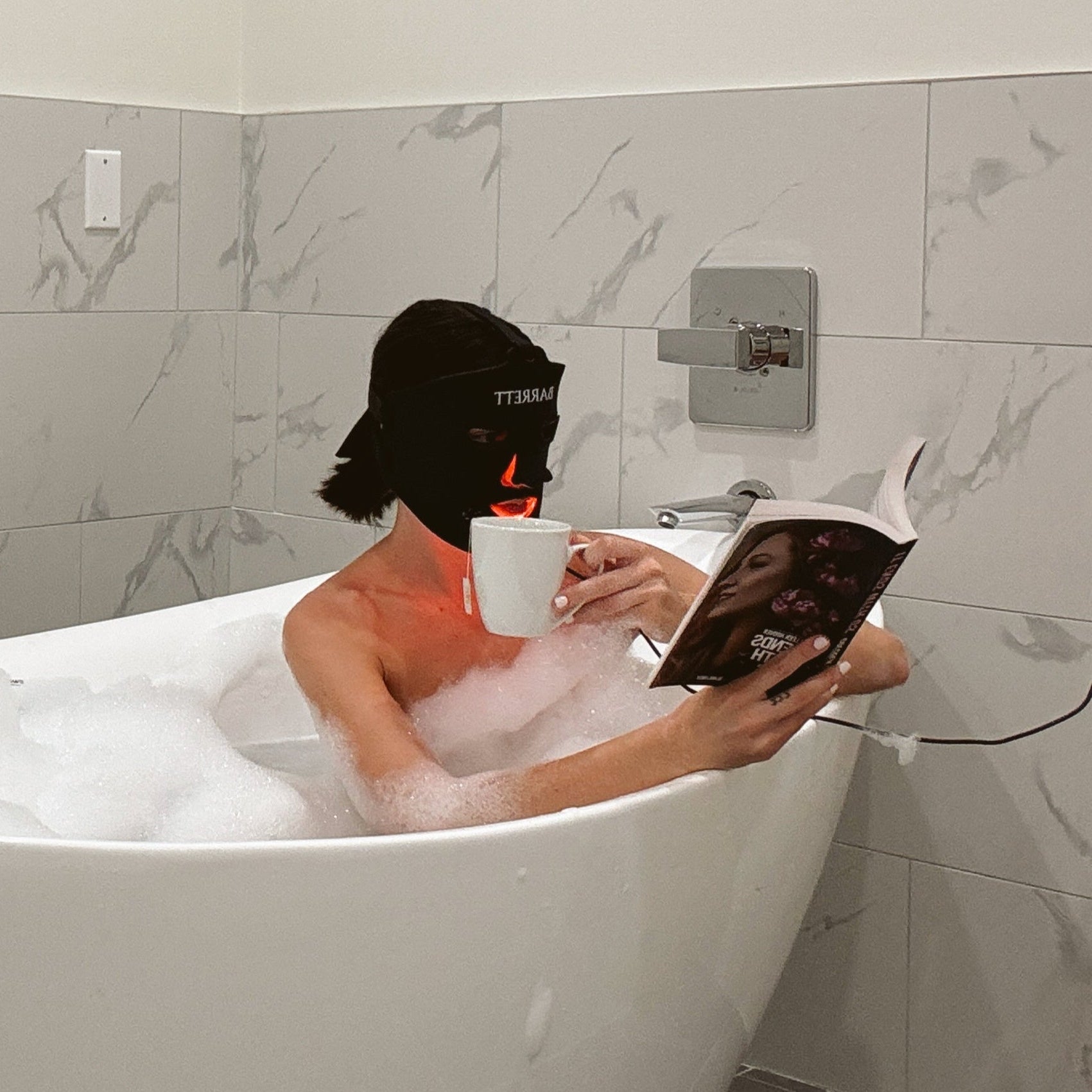 Woman in bath using barrett red light mask and reading while drinking tea
